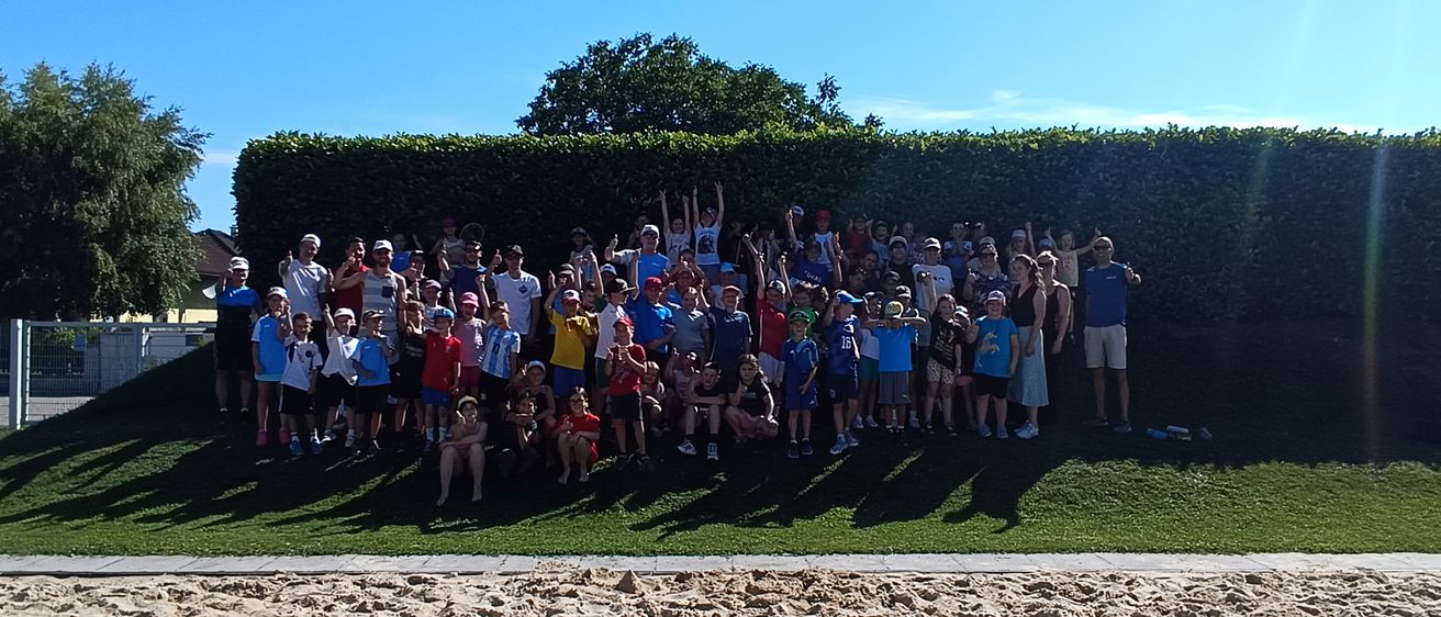 Eine große Gruppe von Menschen, wahrscheinlich ein Sportteam, posiert für ein Foto auf einem Rasenfeld mit einem sandigen Bereich und einem Hecke im Hintergrund.