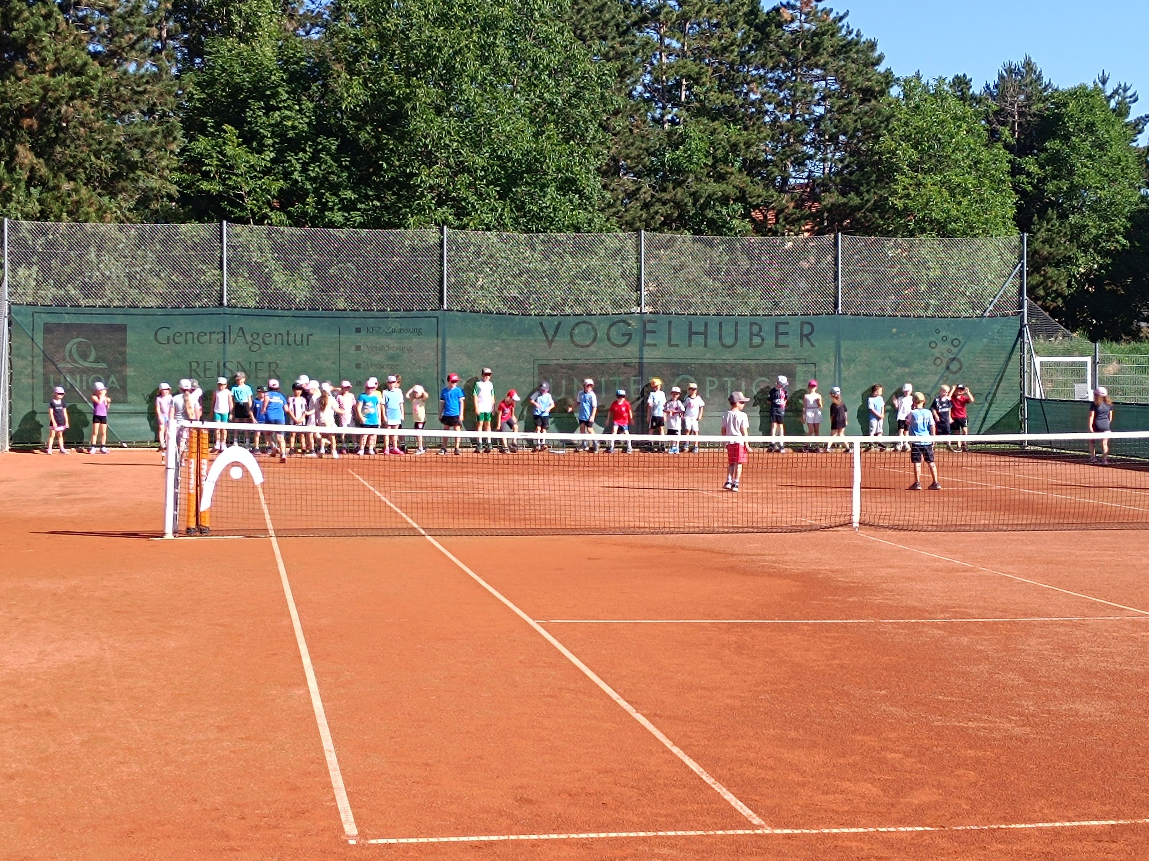 Eine Gruppe von Kindern beobachtet ein Tennisspiel auf einem Sandplatz mit einem Zaun und Bäumen im Hintergrund.
