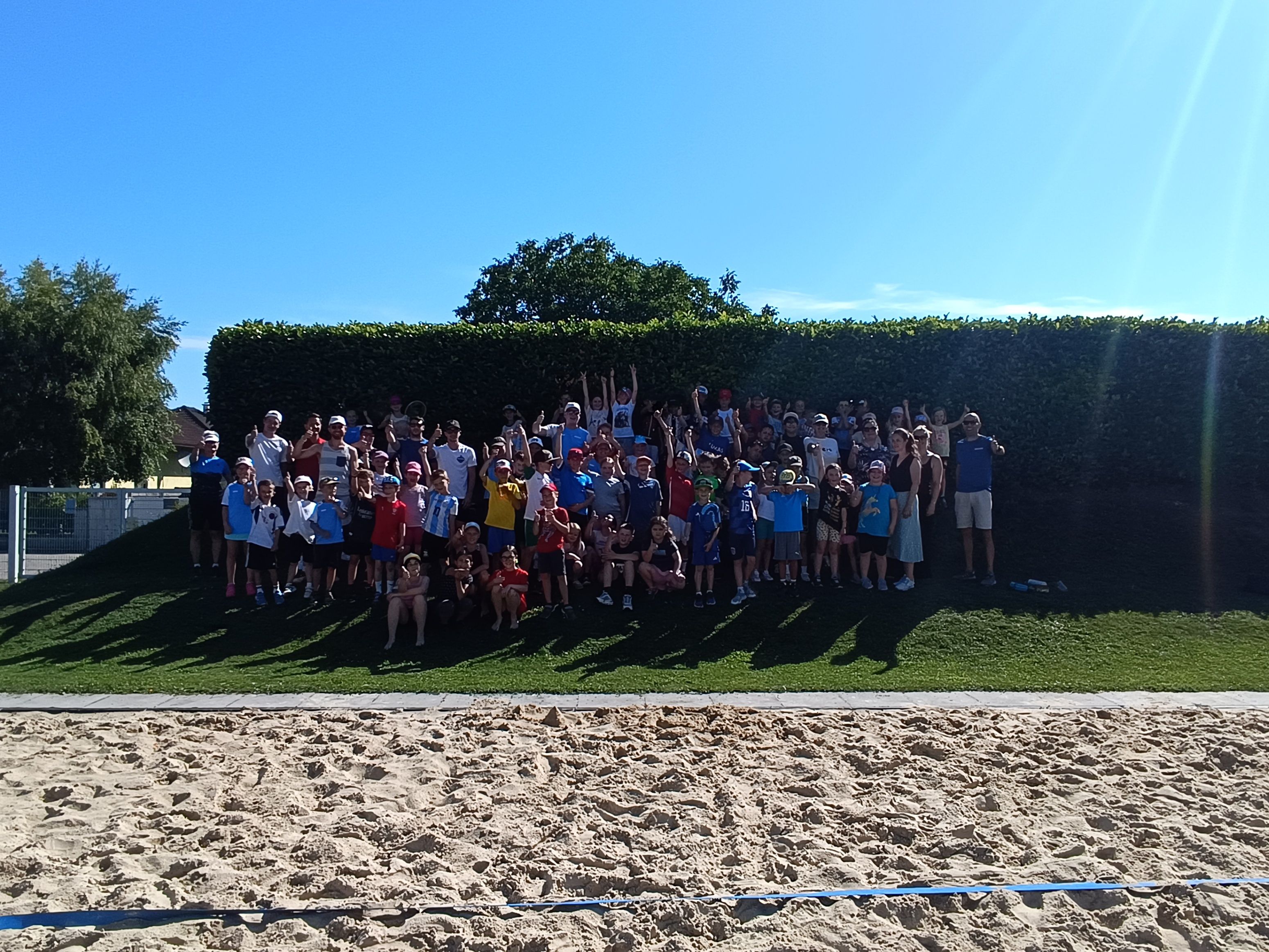 Eine große Gruppe von Menschen, wahrscheinlich ein Sportteam, posiert für ein Foto auf einem Rasenfeld mit einem sandigen Bereich und einem Hecke im Hintergrund.