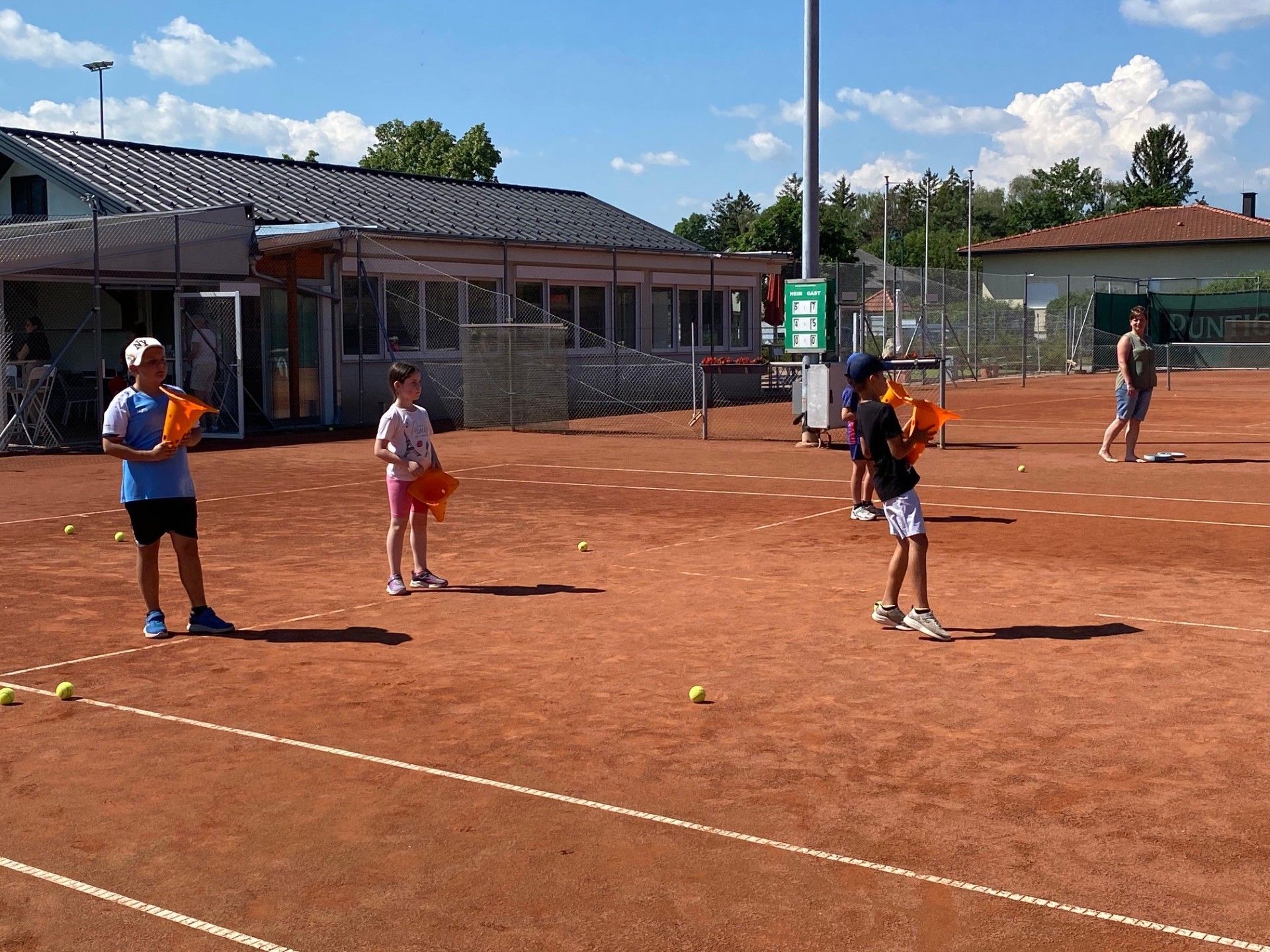Eine Gruppe von Kindern spielt Tennis auf einem Sandplatz. Sie halten Tennisschläger und es liegen Tennisbälle auf dem Boden.