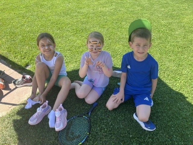 Drei Kinder sitzen auf dem Rasen, zwei Mädchen mit Katzengesichtsbemalung und ein Junge mit grünem Hut. Sie lächeln und posieren für ein Foto mit einem Tennisschläger vor ihnen.