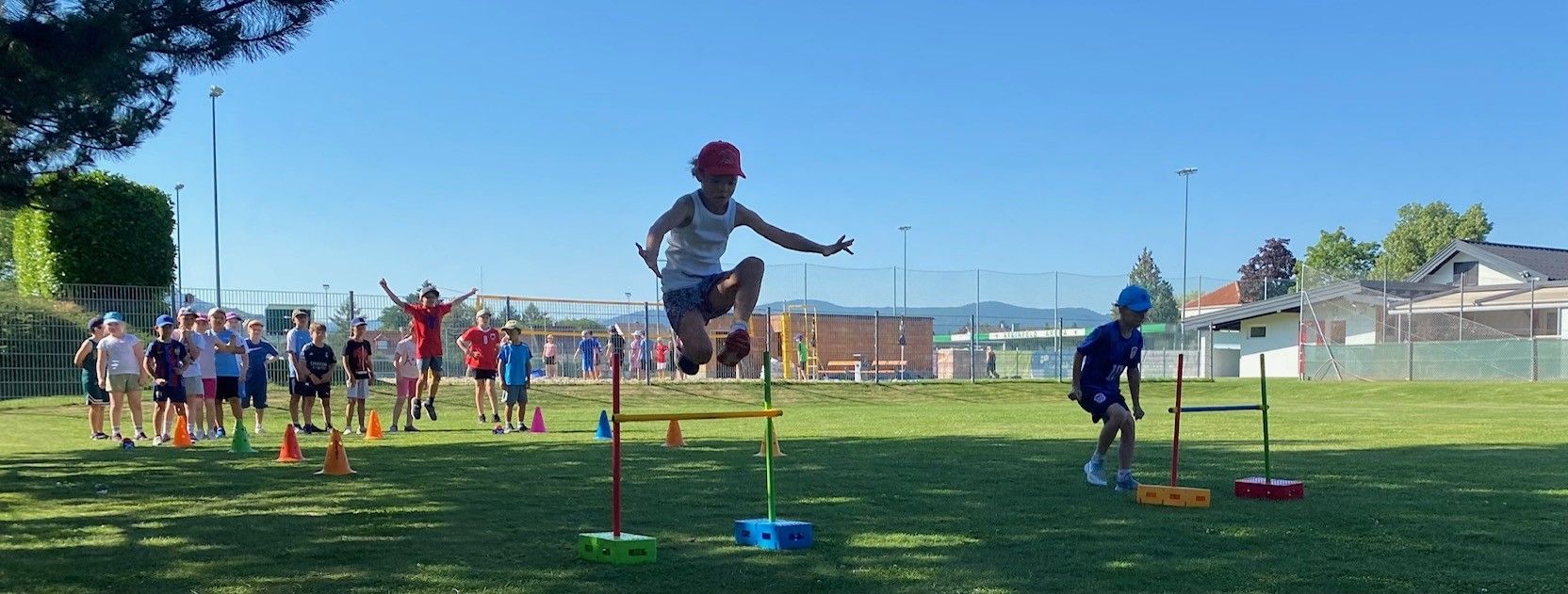 Ein junger Junge springt über eine Hürde auf einem Sportplatz, trägt eine rote Kappe und Turnschuhe. Andere Kinder spielen im Hintergrund.