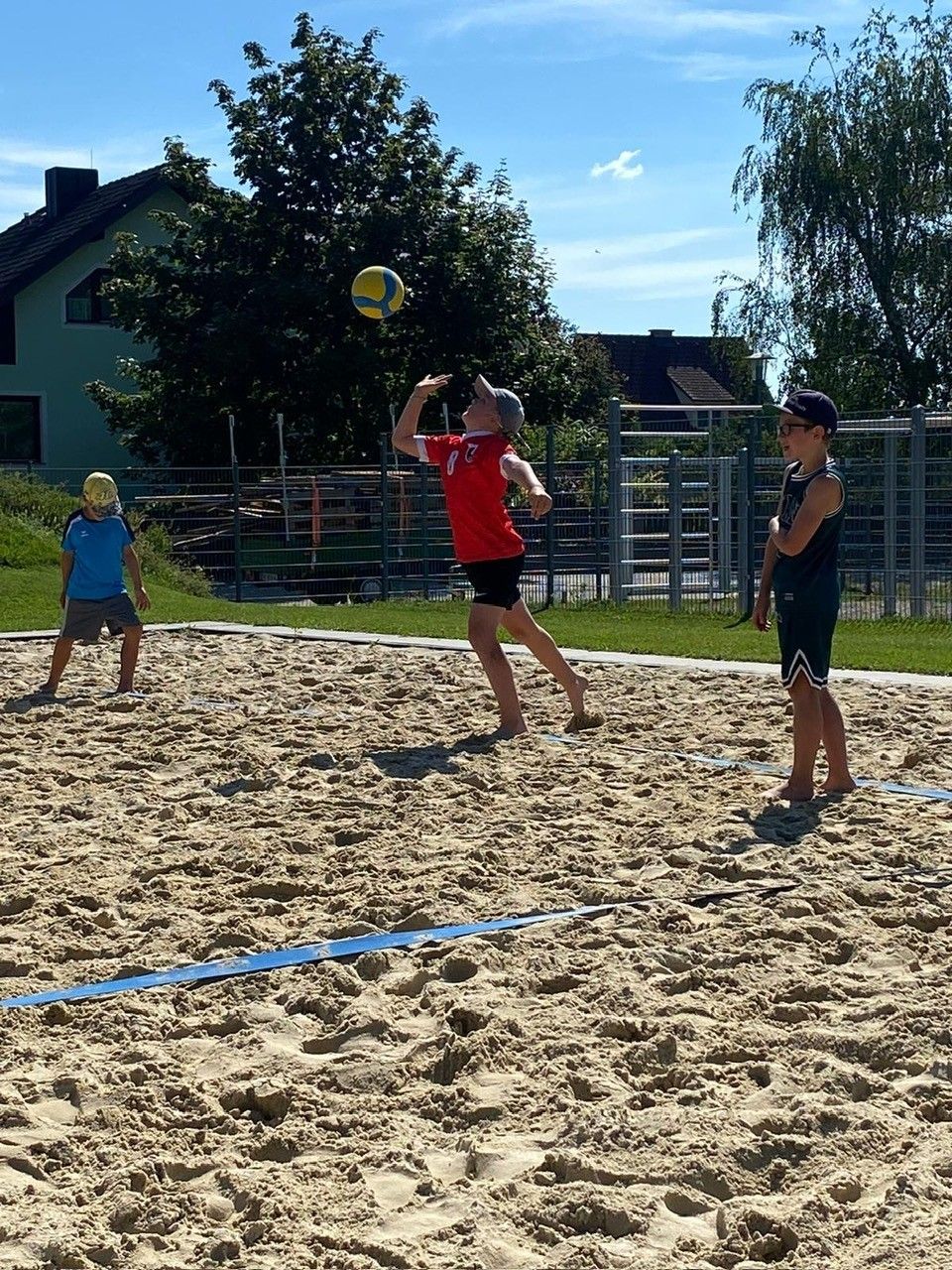 Drei Kinder spielen Volleyball auf einem Sandplatz. Ein Kind in Rot springt, um den Ball zu schlagen.