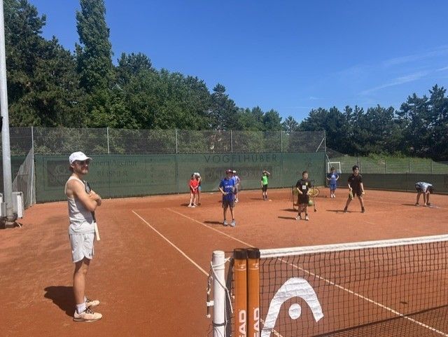 Eine Gruppe von Kindern und einem Mann spielen Tennis auf einem Platz mit Netz und Zaun. Bäume und ein blauer Himmel sind im Hintergrund.