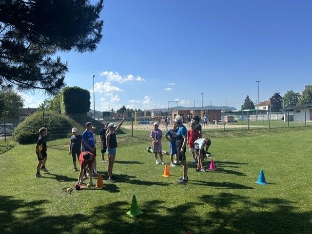 Eine Gruppe von Menschen versammelt sich auf einem Rasenfeld, wahrscheinlich für eine Sportveranstaltung. Sie tragen Mützen und Turnschuhe, und einige halten Tennisschläger. Es gibt orangefarbene und blaue Kegel auf dem Boden, und eine Person beugt sich nach unten. In der Ferne befindet sich ein Zaun mit Netz und ein Gebäude.