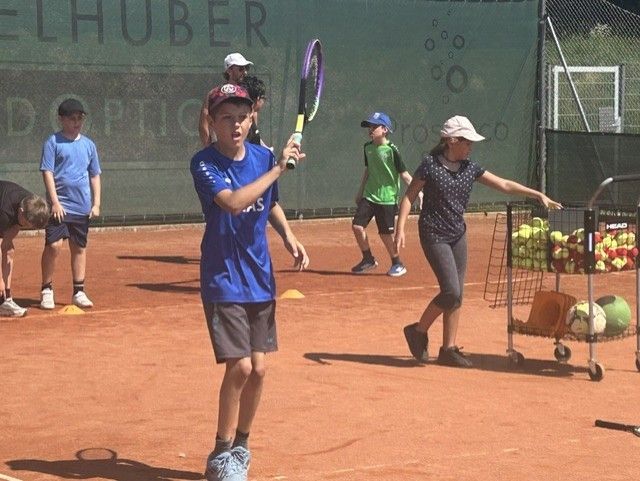 Mehrere Kinder üben Tennis auf einem Sandplatz. Ein Junge in einem blauen Shirt hält einen Tennisschläger, während andere in der Nähe sind, einige beugen sich. Eine Frau hebt Tennisbälle von einem Wagen auf.