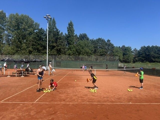 Eine Gruppe von Menschen spielt Tennis auf einem Sandplatz. Einige heben Tennisbälle auf, während andere stehen und zusehen.