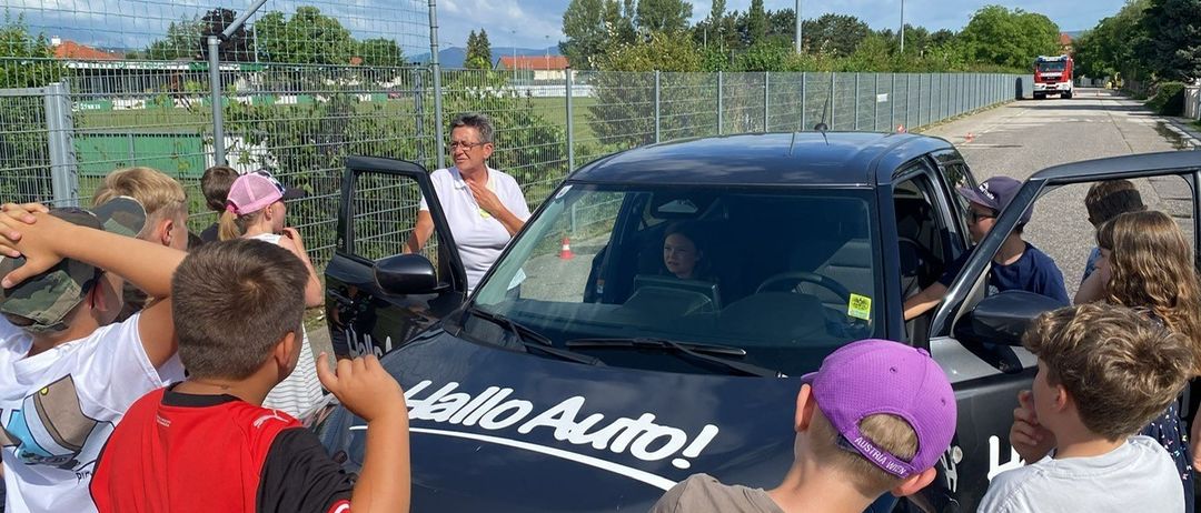 Eine Frau steht neben einem schwarzen Auto mit dem Text 'Hallo Auto!' auf der Motorhaube, während eine Frau im Inneren sitzt und eine Gruppe von Kindern zusieht.