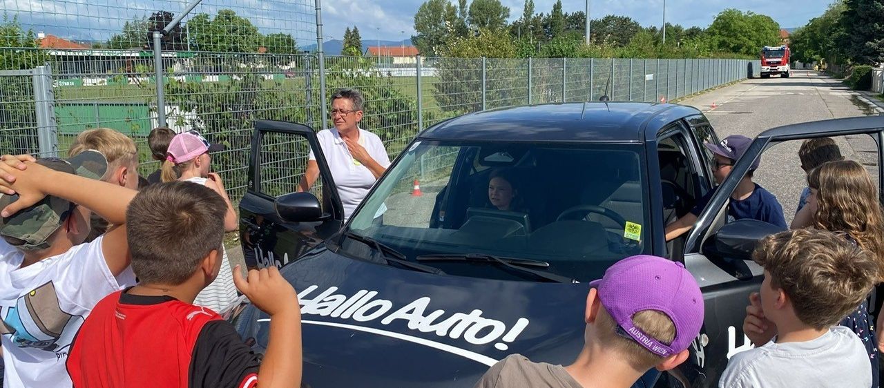 Eine Frau steht neben einem schwarzen Auto mit dem Text 'Hallo Auto!' auf der Motorhaube, während eine Frau im Inneren sitzt und eine Gruppe von Kindern zusieht.