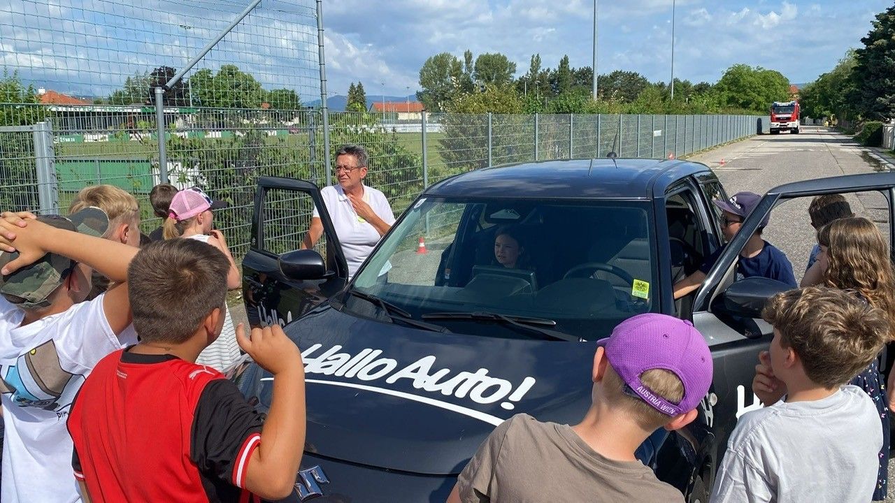 Eine Frau steht neben einem schwarzen Auto mit dem Wort Hello Auto auf der Motorhaube. Eine Frau sitzt auf dem Fahrersitz. Ein Junge und ein Mädchen stehen vor dem Auto.