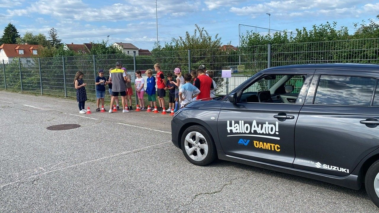Eine Gruppe von Kindern und Erwachsenen steht hinter orangefarbenen Verkehrskegeln vor einem Auto mit dem Text 'Hallo Auto!' an der Seite.