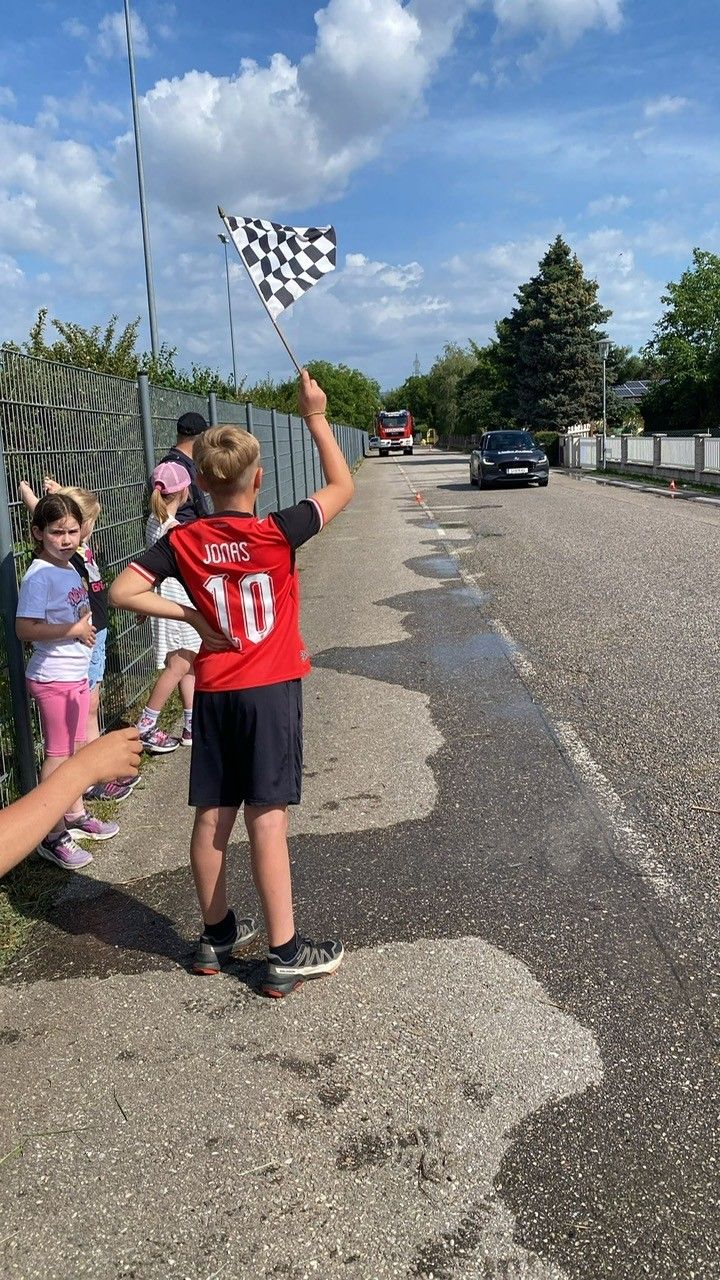 Ein Junge winkt mit einer Fahne mit Schachbrettmuster, während er am Straßenrand steht, mit Kindern und einem Auto im Hintergrund.