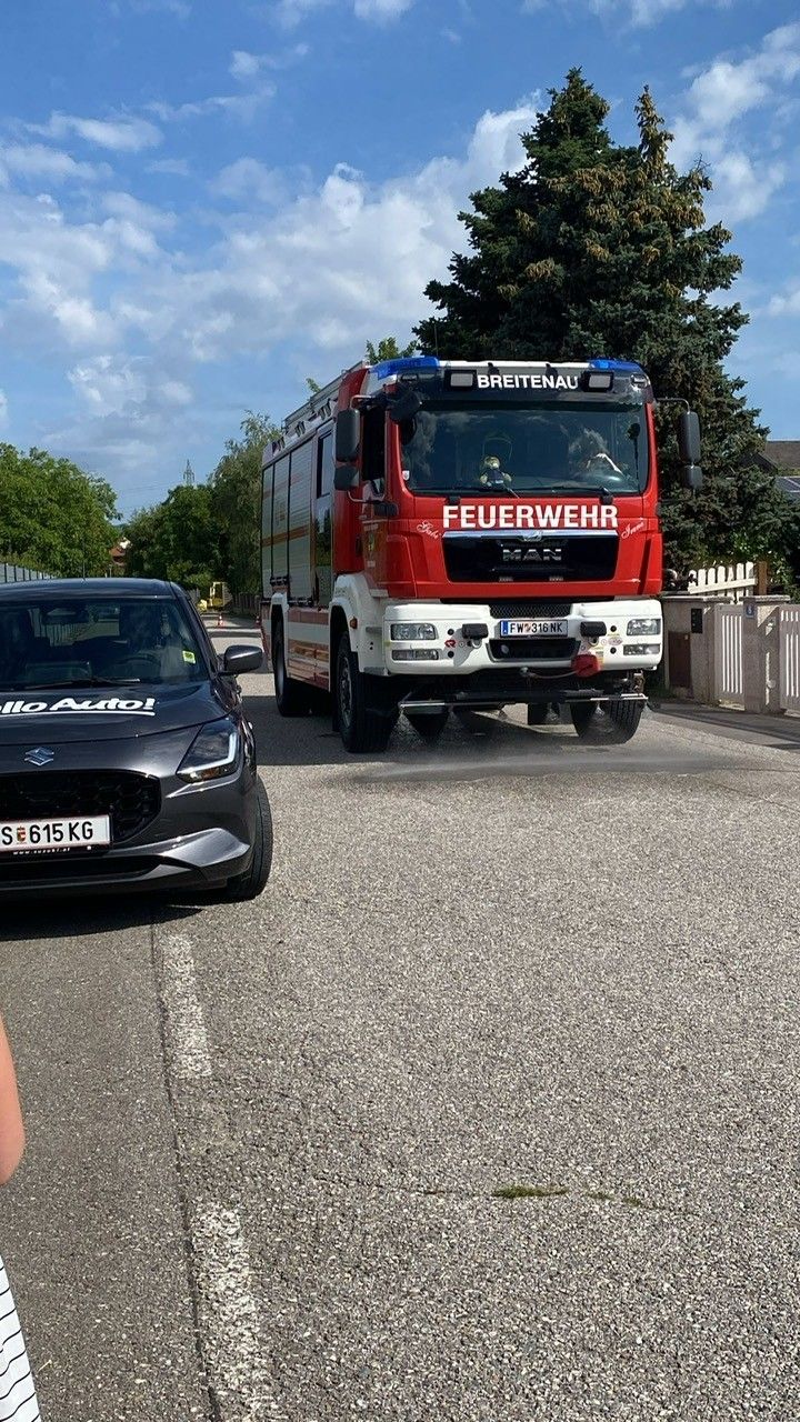Ein roter Feuerwehrwagen mit der Aufschrift 'Feuerwehr' auf der Front fährt auf der Straße. Ein schwarzer Wagen mit dem Kennzeichen 'S 615 KG' ist am Straßenrand geparkt.