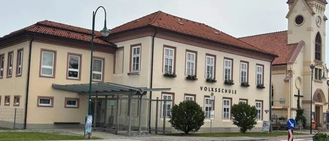 Ein großes Gebäude mit rotem Dach und mehreren Fenstern, beschriftet mit 'Volksschule', befindet sich neben einer Kirche mit Turm und Uhr. Ein Straßenlicht und eine kleine Bushaltestelle sind davor.