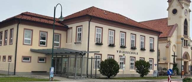 Ein großes Gebäude mit rotem Dach und mehreren Fenstern, beschriftet mit 'Volksschule', befindet sich neben einer Kirche mit Turm und Uhr. Ein Straßenlicht und eine kleine Bushaltestelle sind davor.
