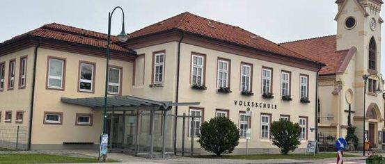 Ein großes Gebäude mit rotem Dach und mehreren Fenstern, beschriftet mit 'Volksschule', befindet sich neben einer Kirche mit Turm und Uhr. Ein Straßenlicht und eine kleine Bushaltestelle sind davor.
