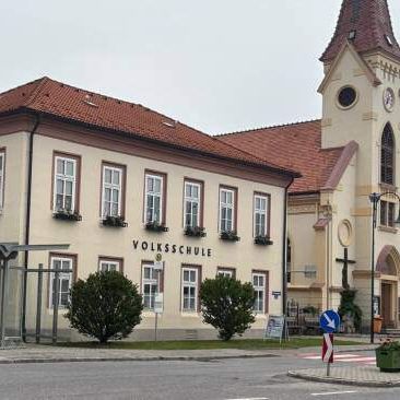 Ein Gebäude mit rotem Dach und mehreren Fenstern steht neben einer Kirche. Das Schild zeigt 'VOLKSSCHULE'. Vor dem Gebäude befindet sich eine Straßenlampe, eine Bushaltestelle und Sträucher.