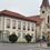 Volksschule Breitenau-Logo. Ein Gebäude mit rotem Dach und mehreren Fenstern steht neben einer Kirche. Das Schild zeigt 'VOLKSSCHULE'. Vor dem Gebäude befindet sich eine Straßenlampe, eine Bushaltestelle und Sträucher.