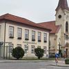 Ein Gebäude mit rotem Dach und mehreren Fenstern steht neben einer Kirche. Das Schild zeigt 'VOLKSSCHULE'. Vor dem Gebäude befindet sich eine Straßenlampe, eine Bushaltestelle und Sträucher.