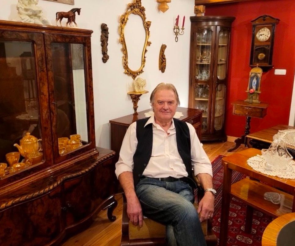 Ein älterer Mann mit grauem Haar und einem Schnurrbart sitzt in einem Stuhl in einem Zimmer voller antiker Möbel. Hinter ihm steht ein hölzerner Schrank mit Glastüren, ein Spiegel und eine Pferdestatue. Auf der rechten Seite befindet sich eine hölzerne Uhr und auf der linken Seite ein hölzerner Schrank mit Teekanne und Tassen.