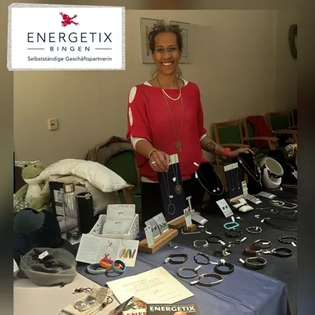 Eine Frau steht hinter einem Tisch mit verschiedenen Schmuckstücken. Der Tisch hat eine blaue Tischdecke und ist mit verschiedenen Armbändern, Halsketten und Ohrringen gefüllt. Auf dem Tisch befinden sich auch Bücher, ein Kissen und ein Glas. Die Frau trägt eine rote Bluse und eine Brille.