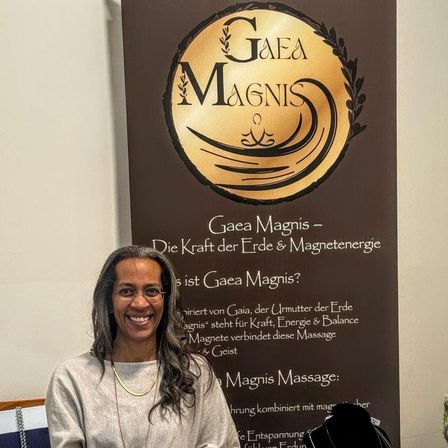 Eine Frau steht hinter einem Tisch mit Halsketten und Armbändern zur Schau. Sie hält ein Weinglas. Hinter ihr ist ein Banner mit einem Logo und Text. Ein grüner Frosch und ein Buch sind auf dem Tisch.