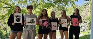 Six young people hold signs in a park under trees, showing drawings and words. A woman smiles with a bracelet on her wrist.