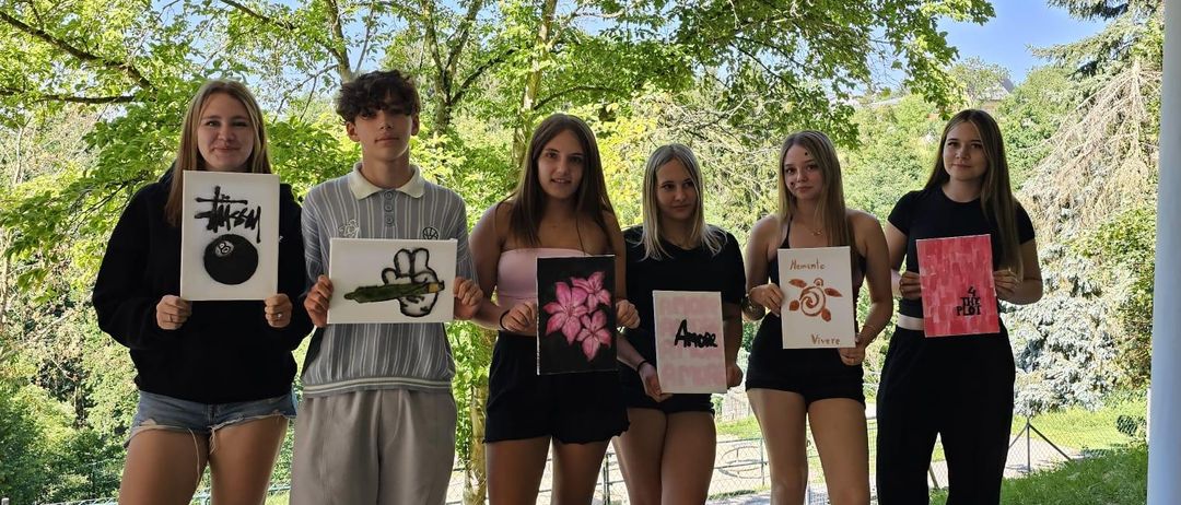 Six young people hold signs in a park under trees, showing drawings and words. A woman smiles with a bracelet on her wrist.