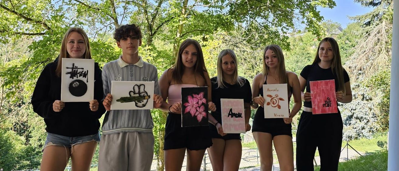 Sechs junge Menschen halten Schilder in einem Park unter Bäumen, mit Zeichnungen und Wörtern. Eine Frau lächelt mit einem Armband an ihrem Handgelenk.