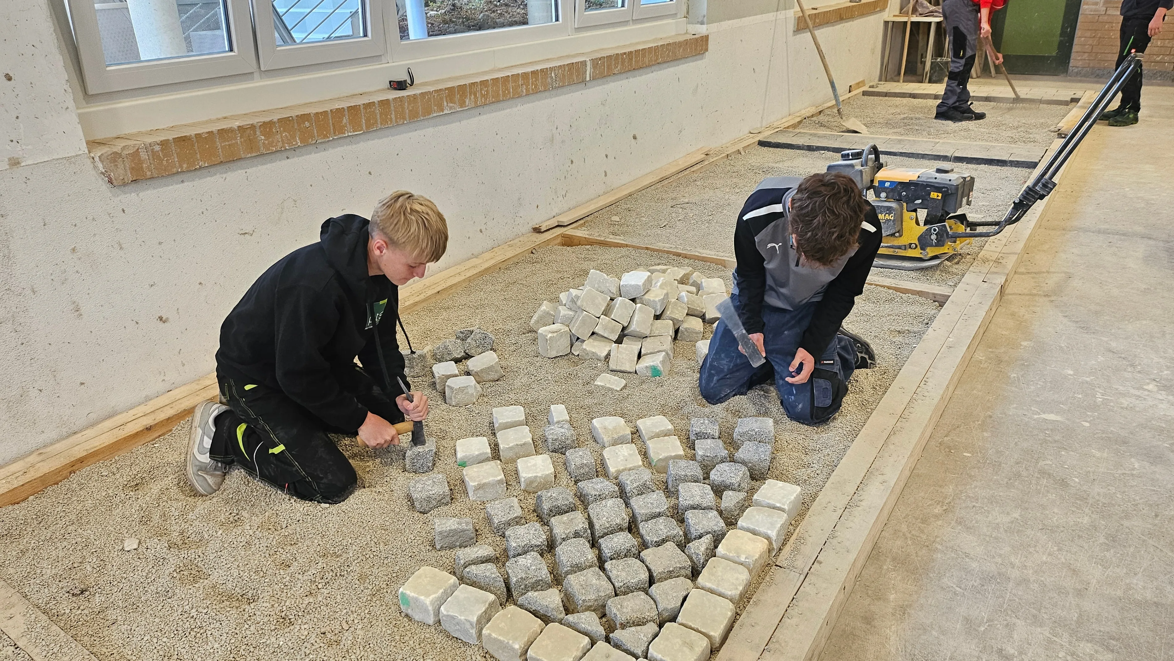 Zwei junge Jungen arbeiten auf einer Baustelle, knien auf dem Boden und ordnen Steine an. Ein Junge hält einen Hammer, und der andere benutzt ein Werkzeug, um die Steine zu formen. Hinter ihnen sind ein großes Fenster und eine Tür zu sehen, und eine Person steht in der Nähe einer Maschine.