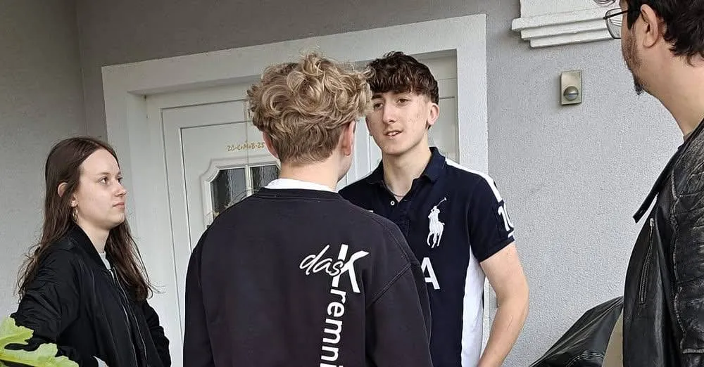 Drei junge Leute stehen vor einem Gebäude. Der Junge in der Mitte trägt ein Polohemd mit der Nummer 10, während der Junge auf der rechten Seite ein schwarzes und weißes Polohemd trägt. Der Junge auf der linken Seite trägt eine schwarze Jacke mit weißem Text.