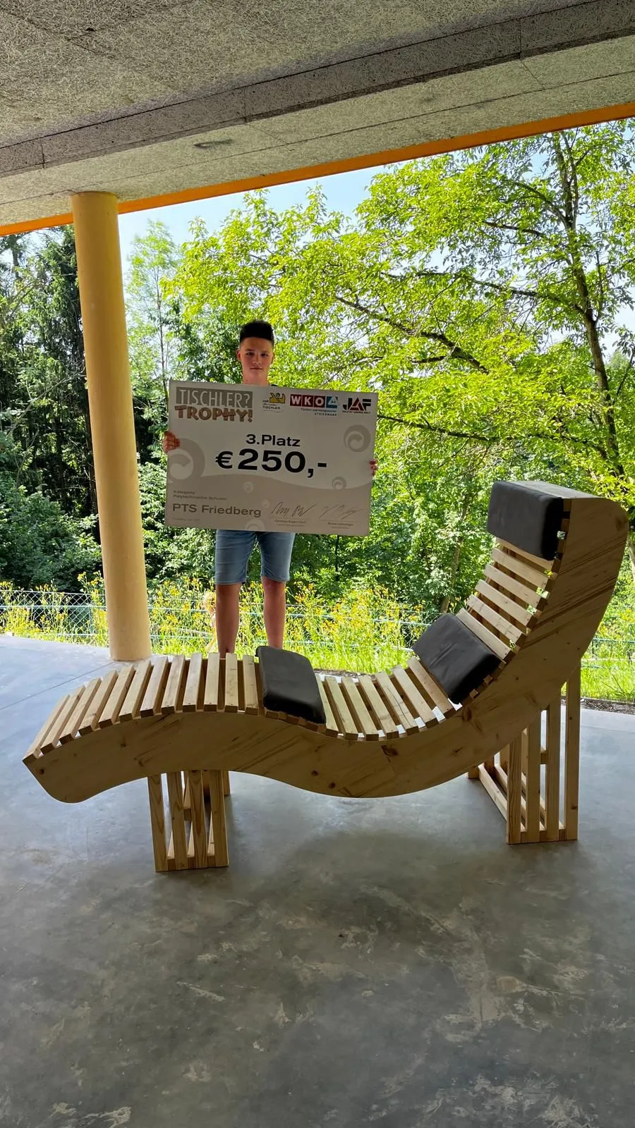 Ein junger Mann steht im Freien und hält eine Trophäe mit der Aufschrift '3. Platz' und '250 Euro'. Er steht auf einem hölzernen Liegestuhl mit schwarzen Kissen.