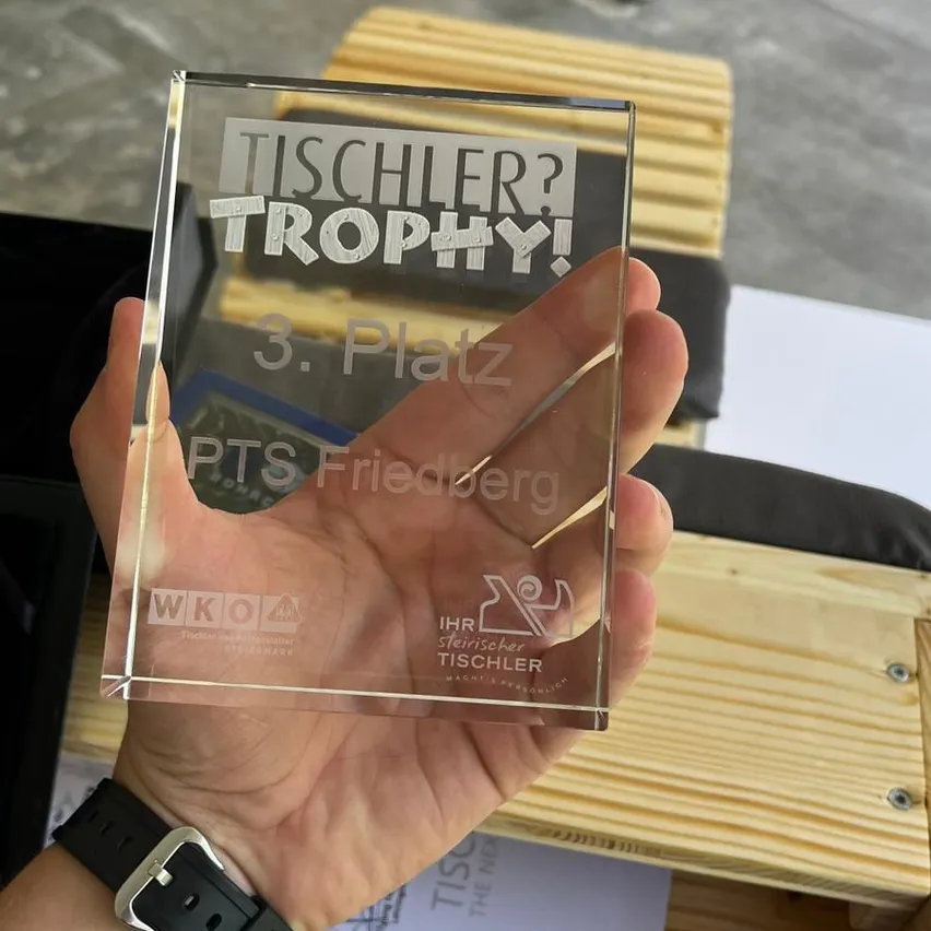 Eine Person hält eine transparente Acryl-Trophäe mit dem Text Tischler? Trophy! 3. Platz PTS Friedberg. Darunter ist das Logo von Tischler gedruckt. Die Trophäe steht auf einer Holzoberfläche.