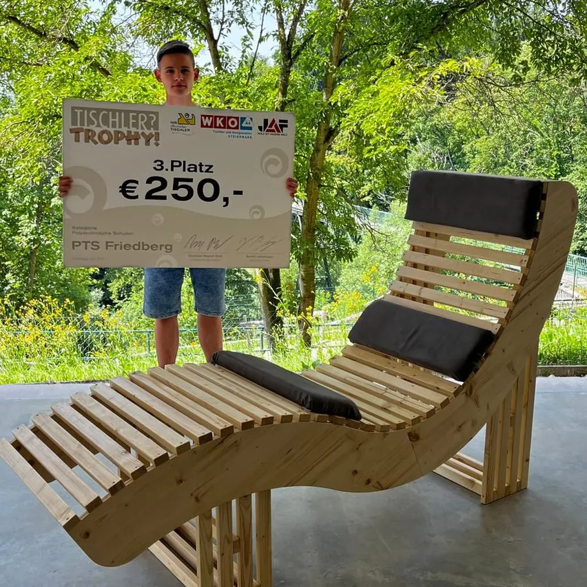 Ein junger Junge hält eine Trophäe, die 3. Platz und 250 Euro zeigt, neben einem gebogenen Holz-Lounge-Sessel in einem Garten.