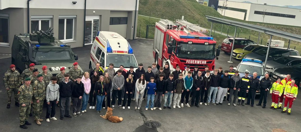 Eine Gruppe von Schülern posiert vor einem Krankenwagen und einem Feuerwehrauto. Ein Hund liegt vor ihnen auf dem Boden.