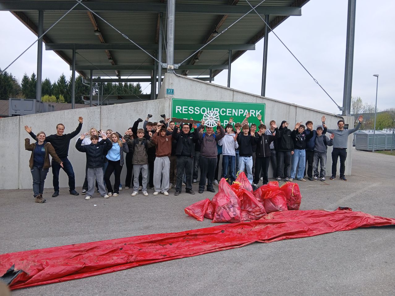 Eine Gruppe von Menschen steht vor einem Gebäude mit einem Schild, das 'Ressourcenpark' besagt. Sie alle lächeln und posieren für ein Foto. Einige halten Plastiktüten. Ein großes rotes Planen liegt auf dem Boden.