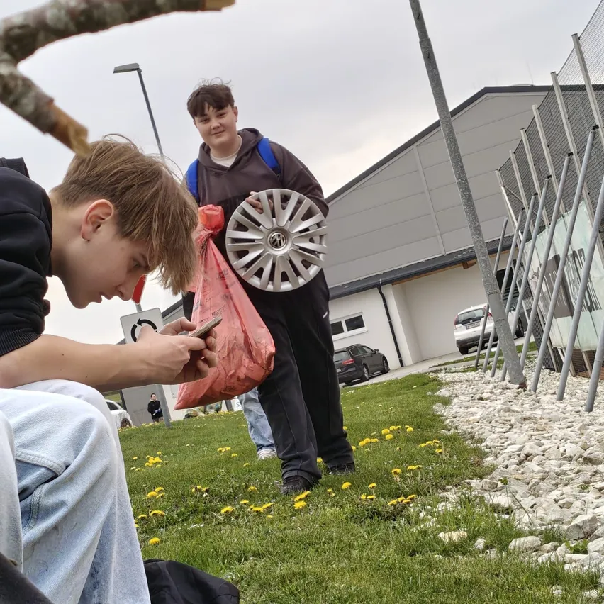 Zwei junge Jungen sitzen auf dem Gras. Ein Junge hält ein Handy und sieht nach unten, während der andere Junge einen roten Plastikbeutel mit einem Rad hält. Dahinter steht ein Gebäude mit einem Zaun.