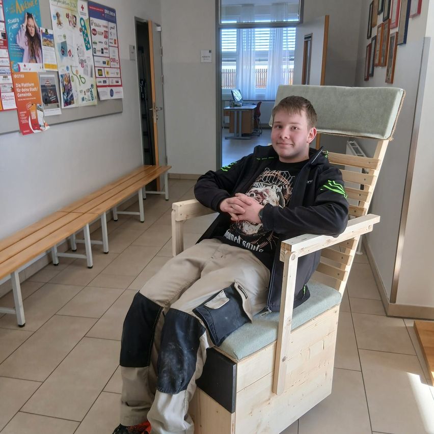 Ein junger Mann sitzt in einem Holzstuhl in einem Flur und lächelt für die Kamera. Dahinter ist eine Pinnwand mit verschiedenen Postern an einer Wand befestigt.