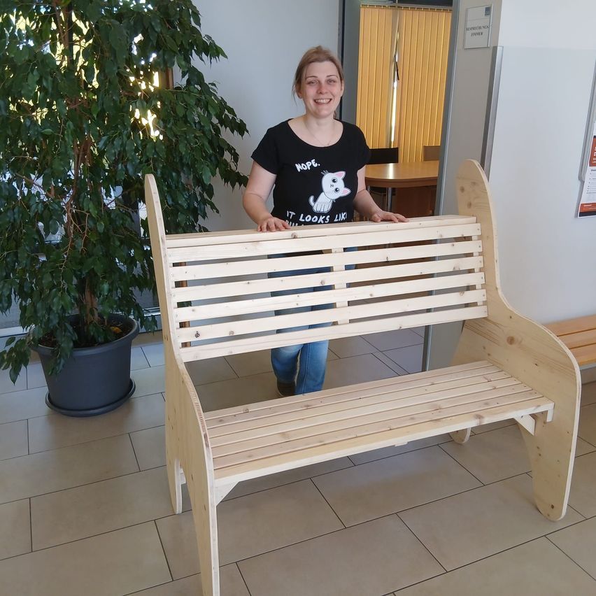 Eine Frau steht hinter einer Holzbank in einem Raum mit einem Topf mit Pflanzen und einem Tisch mit Stühlen.