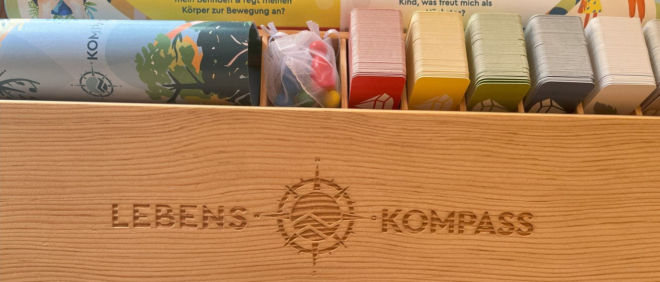 Eine Holzkiste zeigt einen Kompass mit der Aufschrift 'Lebens-Kompass', mit verschiedenen Karten, bunten Kugeln und Papierrollen im Inneren. Zwei Rollen haben gedruckte Botschaften auf ihnen.