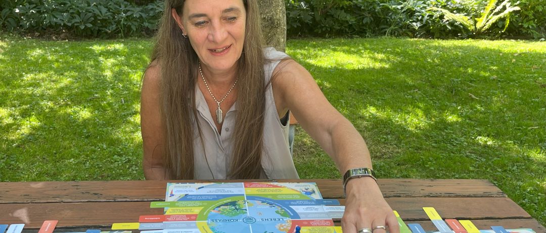 A woman sits at a picnic table in a garden, playing a board game. The board has colored pieces and maps. She wears a necklace and rings.