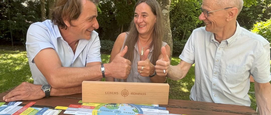 Three people sit at a table outside, smiling. A wooden box labeled Lebens-Kompass sits on the table in front of them. The box contains a board game with colored pieces and cards.