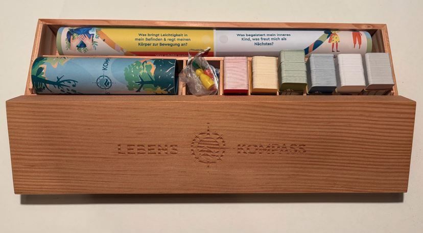 A wooden box contains a compass kit with various cards, a booklet, and small objects. The box is labeled 'Lebenskompass'.