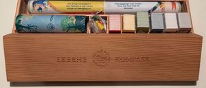 Eine Holzkiste enthält mehrere Gegenstände, darunter eine gelbe und weiße Rolle, eine blaue Rolle, einen durchsichtigen Plastikbeutel und mehrere Karten.