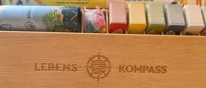 Eine Holzkiste enthält mehrere Gegenstände, darunter Karten, Papierrollen und kleine Tüten, die mit Text auf Deutsch beschriftet sind. Die Kiste selbst hat einen Kompass und die Wörter 'Lebenskompass' eingraviert.