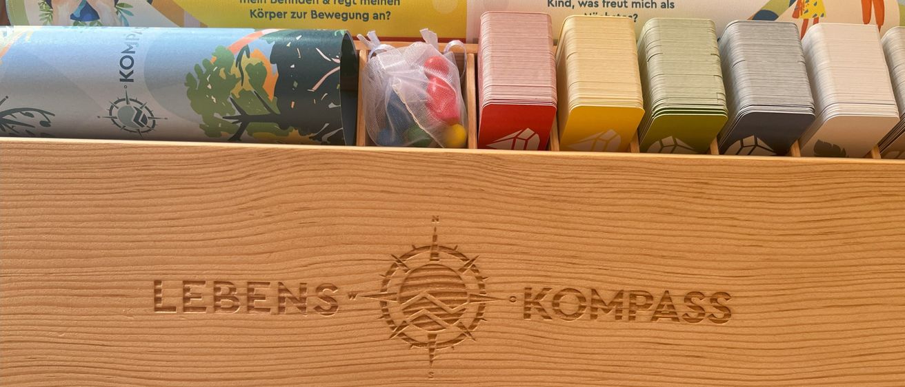 Eine Holzkiste enthält mehrere Gegenstände, darunter Karten, Papierrollen und kleine Tüten, die mit Text auf Deutsch beschriftet sind. Die Kiste selbst hat einen Kompass und die Wörter 'Lebenskompass' eingraviert.