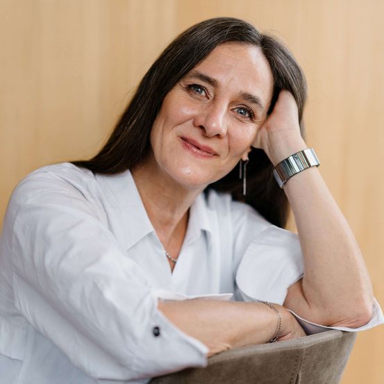 Eine Frau lächelt und schaut direkt in die Kamera. Sie hat ihre rechte Hand an der Wange und ihr linker Arm ruht auf einem Stuhl.