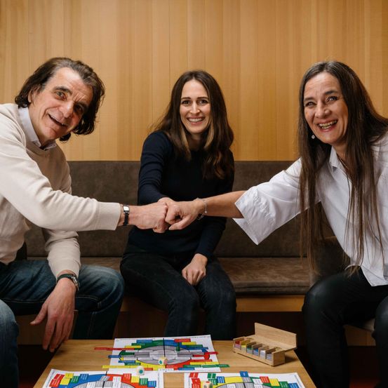 Drei Personen sitzen auf einer Couch in einem Studio. Sie schütteln sich die Hände und lächeln. Ein farbenfrohes Brettspiel liegt auf dem Tisch vor ihnen.