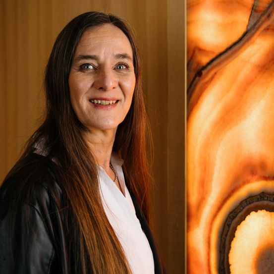 Eine lächelnde Frau mit langen braunen Haaren steht vor einer Wand mit einem welligen orange-braunen Muster und trägt eine schwarze Jacke und ein weißes Hemd.