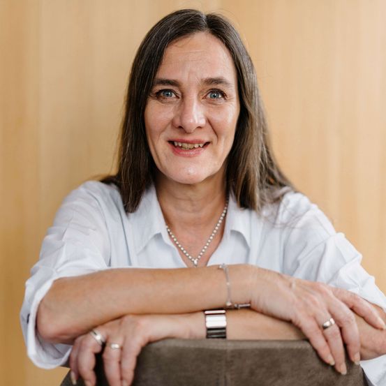 Porträt einer lächelnden Frau mit braunen Haaren, die ein weißes Hemd, eine Halskette und eine Uhr trägt, mit verschrünkten Armen auf einem Holzstuhl sitzt.